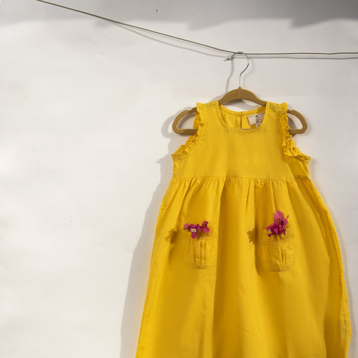 A sunshine yellow colored sleeveless dress for kids in cotton mulmul. This mid length dress has front pockets and a back button fastenings, accentuated with ruffles on the sleeve, neck and pockets.