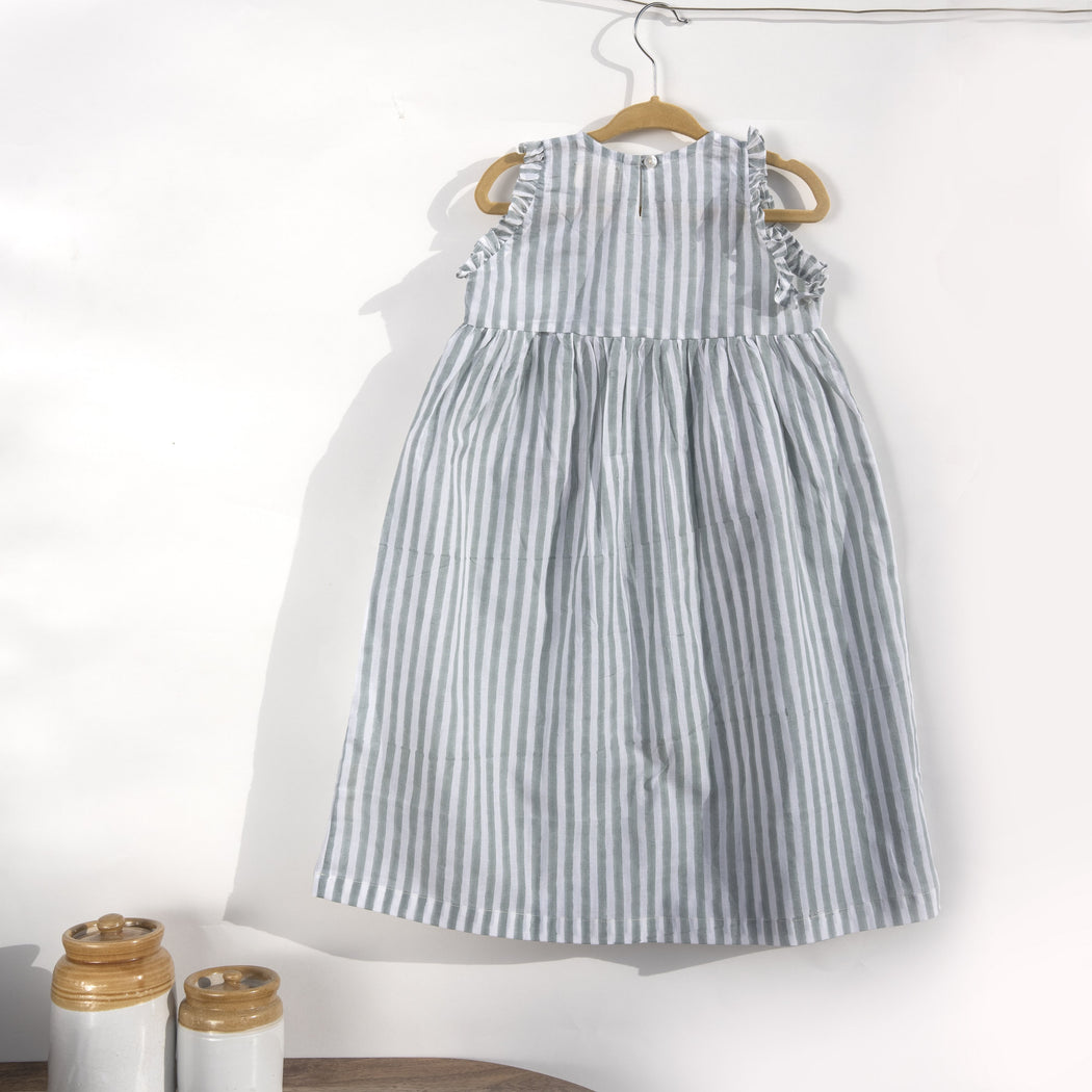 A white cotton mulmul dress for kids with hand-block printed pigeon grey stripes. This mid length dress has front pockets and a back button fastenings accentuated with ruffles on the sleeve, neck and pockets.