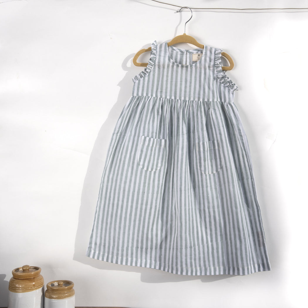 A white cotton mulmul dress for kids with hand-block printed pigeon grey stripes. This mid length dress has front pockets and a back button fastenings accentuated with ruffles on the sleeve, neck and pockets.