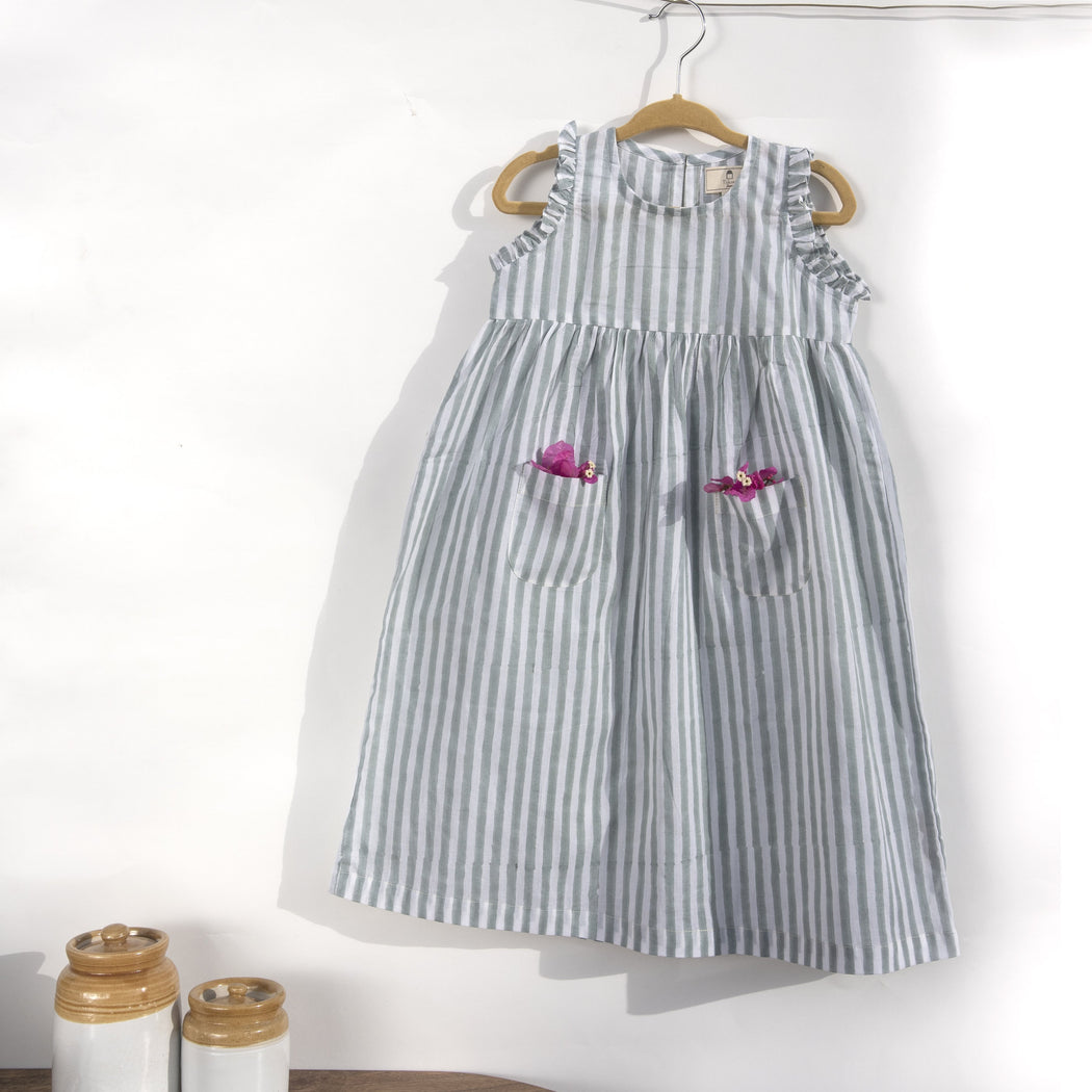 A white cotton mulmul dress for kids with hand-block printed pigeon grey stripes. This mid length dress has front pockets and a back button fastenings accentuated with ruffles on the sleeve, neck and pockets.